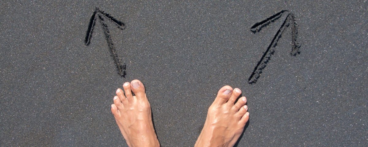 Dos pies desnudos sobre la arena. Delante de cada pie hay una flecha que indica como dos caminos distintos, dos direcciones hacia las que esta persona se podría dirigir.