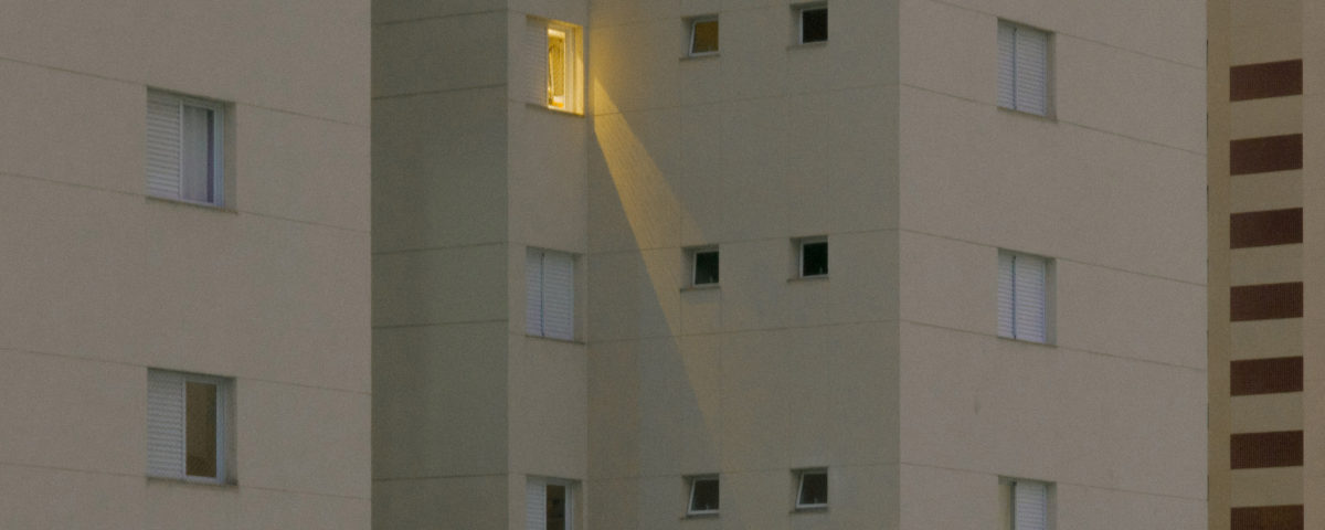 Vista de la fachada de un gran edificio de pisos en el que se ven numerosas ventanas, pero solo una con la luz encendida.